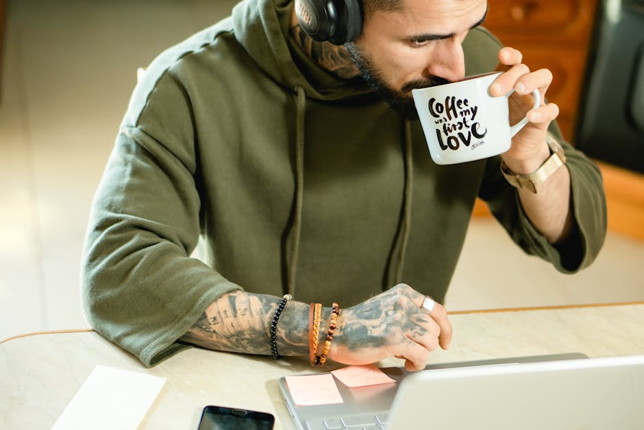Tattooed man in a hoodie drinks coffee while working on a laptop indoors. Lifestyle and leisure concept.